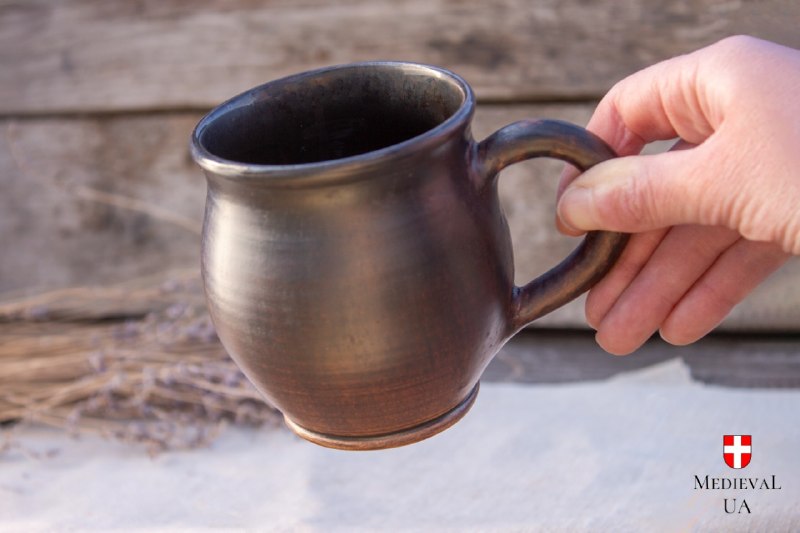 Hand holding a dark milk-fired medieval ceramic tea mug with a rounded belly and glazed interior