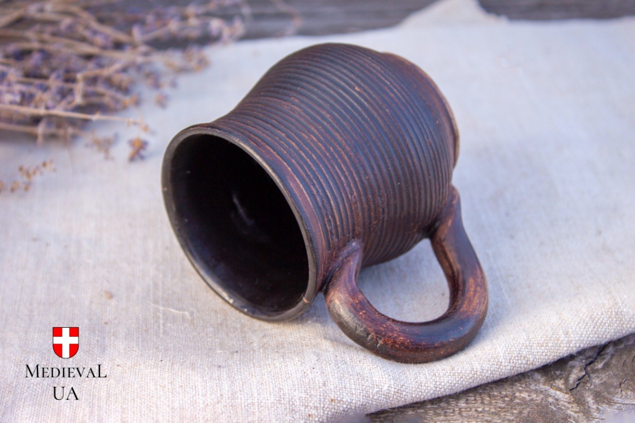 Small dark milk-fired ribbed clay mug resting on linen, showing the looped handle and ridged body