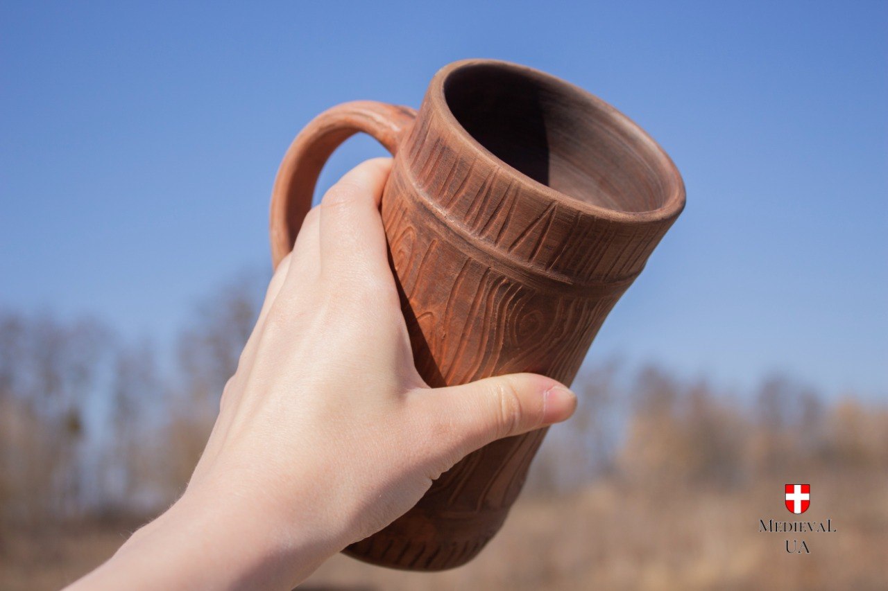 Hand holding the red clay medieval mug up to the sky, showing the carved texture and unglazed interior