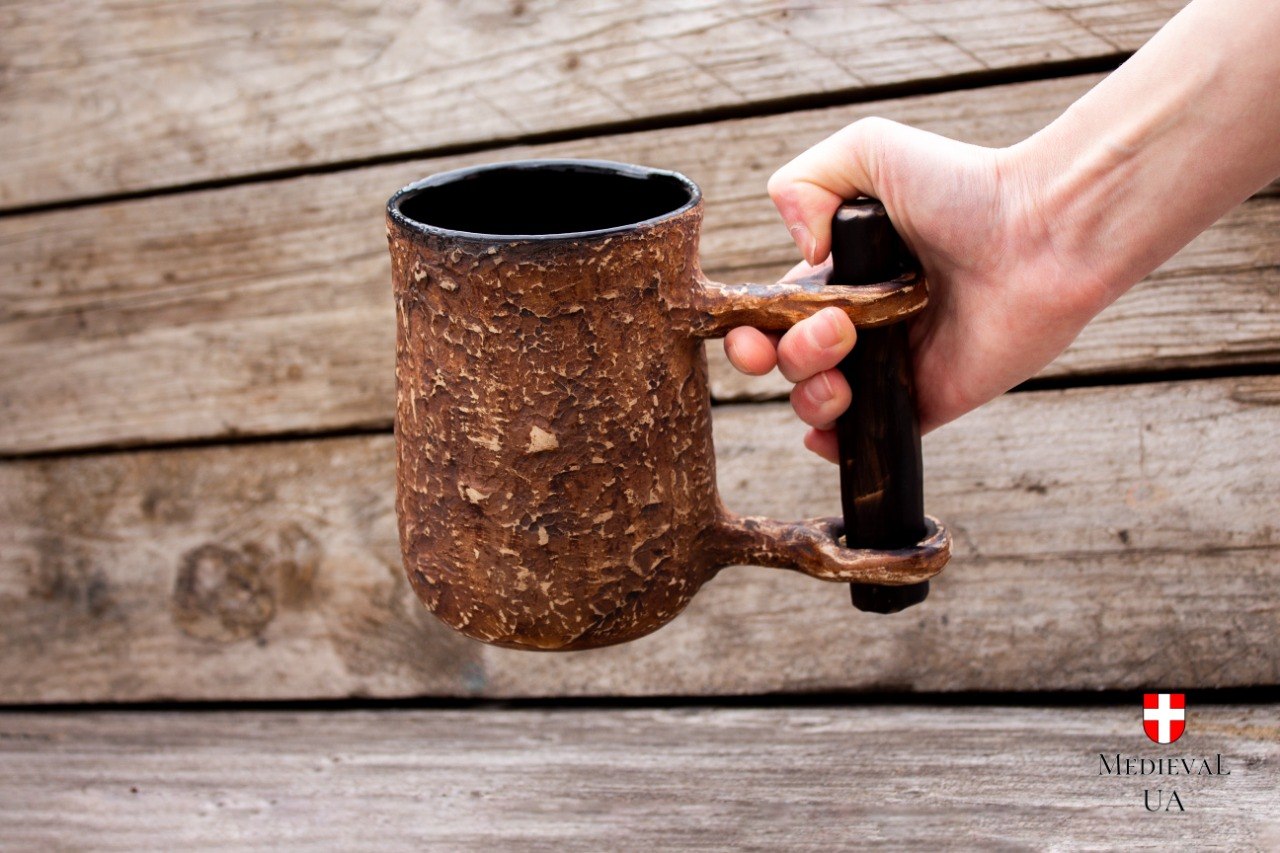 Hand holding the medieval ceramic tankard by its dark wood-like crossbar grip against a wooden wall