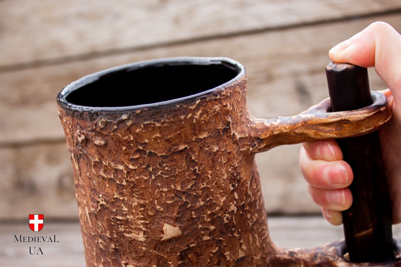Handmade milk-fired medieval beer tankard held by its wooden crossbar handle, showing the textured amber body and black glossy glazed interior
