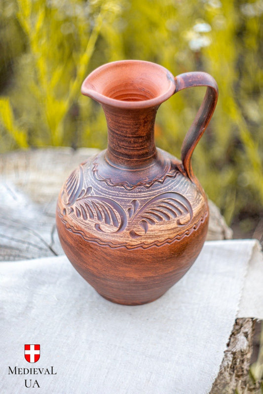 Upright red clay pitcher with carved leaf and ribbon motif and looped handle on linen