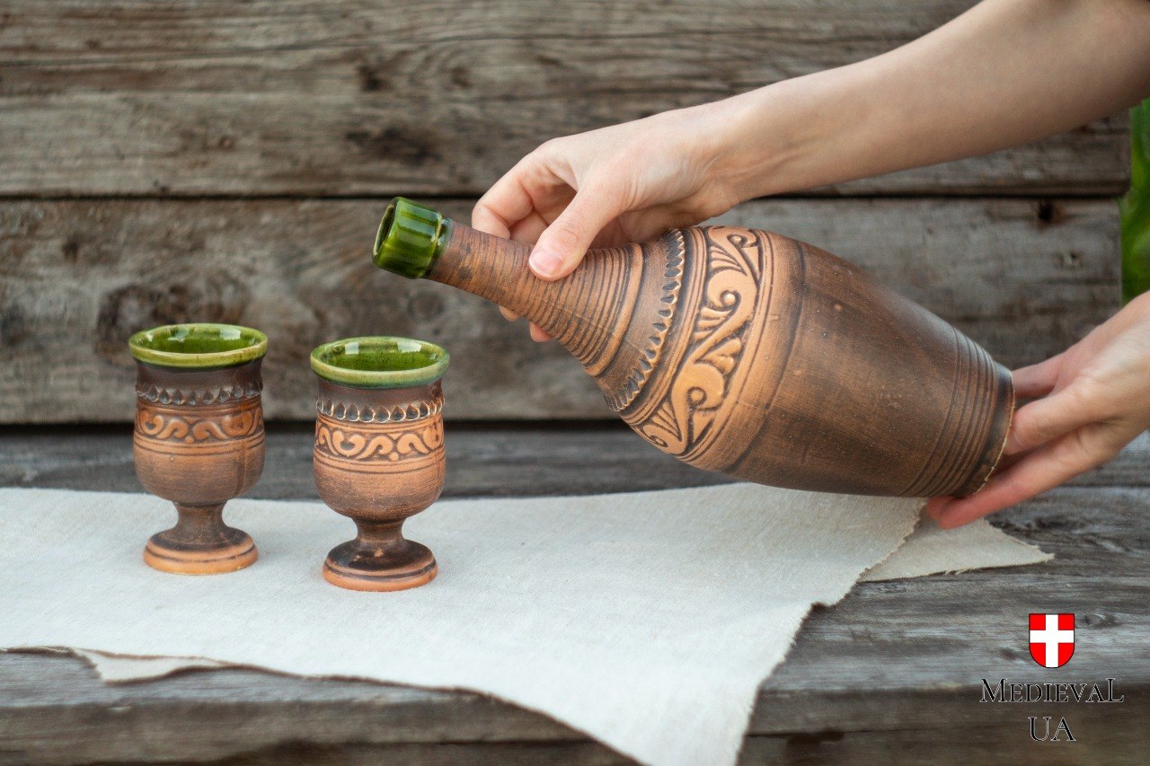 Ceramic Celtic mead bottle tilted to pour beside two matching green-glazed goblets on linen