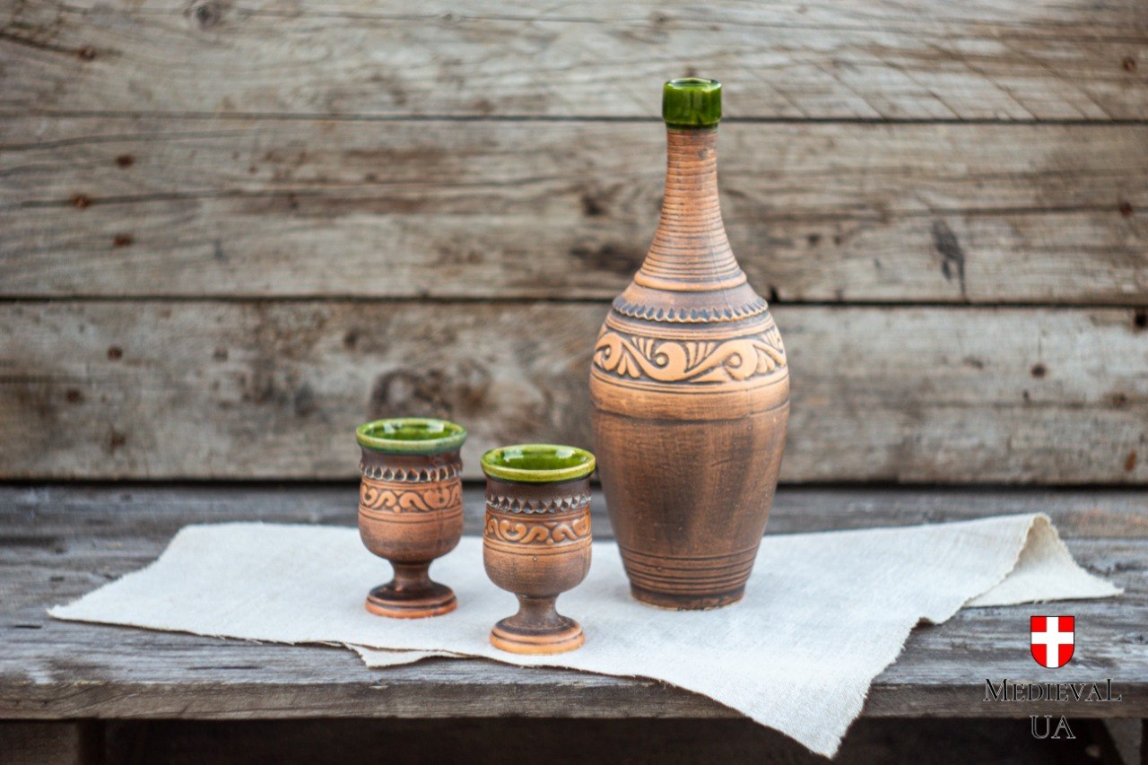 Complete Celtic mead set with clay bottle and two green-glazed goblets on linen and rustic wood