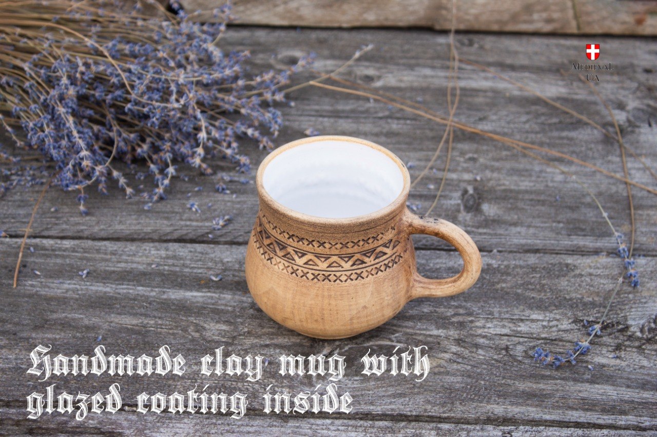 Handmade tan clay medieval mug with an engraved geometric band and white glazed interior on weathered wood with lavender
