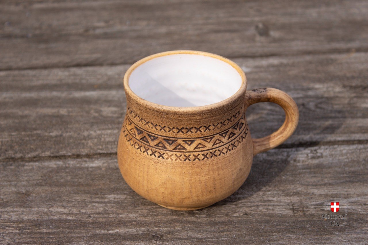 Light tan rustic clay mug with a carved folk pattern and white glaze inside, three-quarter view