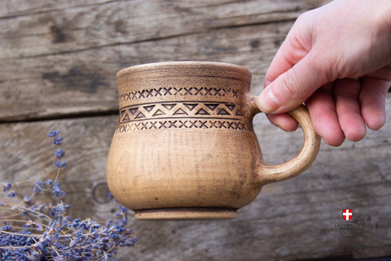 Hand holding the engraved tan clay medieval mug, showing the carved band and footed base