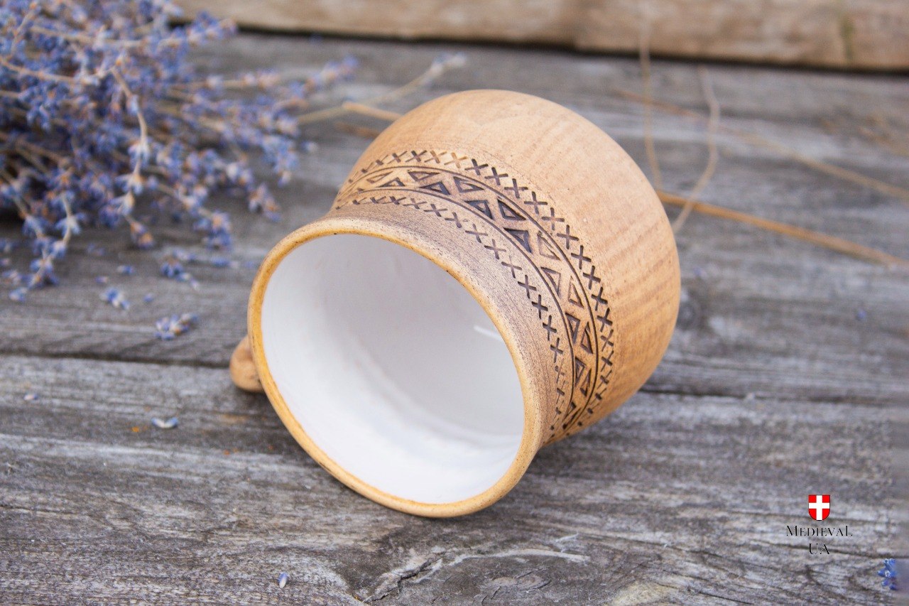 Tan clay mug tipped on its side showing the white glazed interior and carved geometric band