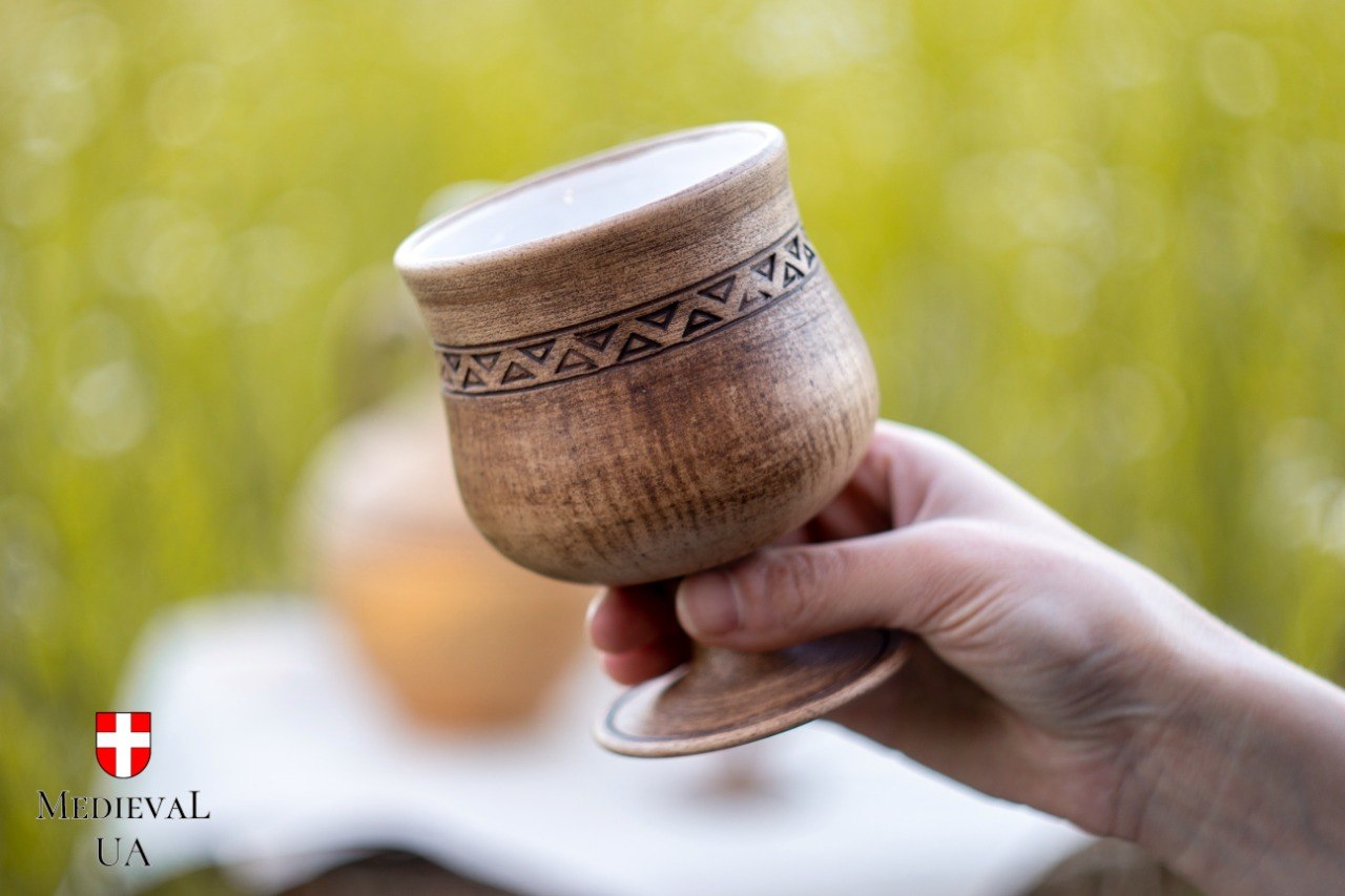 Hand holding a handmade ceramic wine goblet with a carved triangle pattern in a sunny green meadow