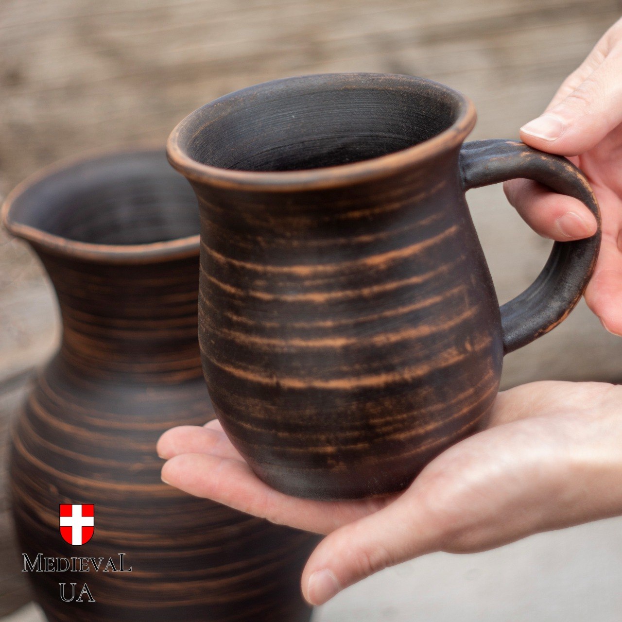 Handmade red clay mug held in hand beside matching jug, showing copper-striped rustic finish
