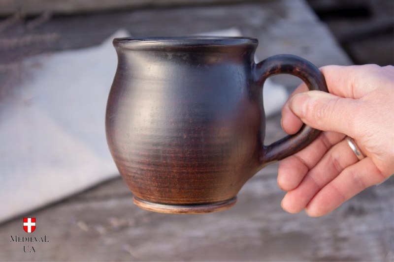 Round-bellied dark brown milk-fired clay mug held in hand against weathered wood