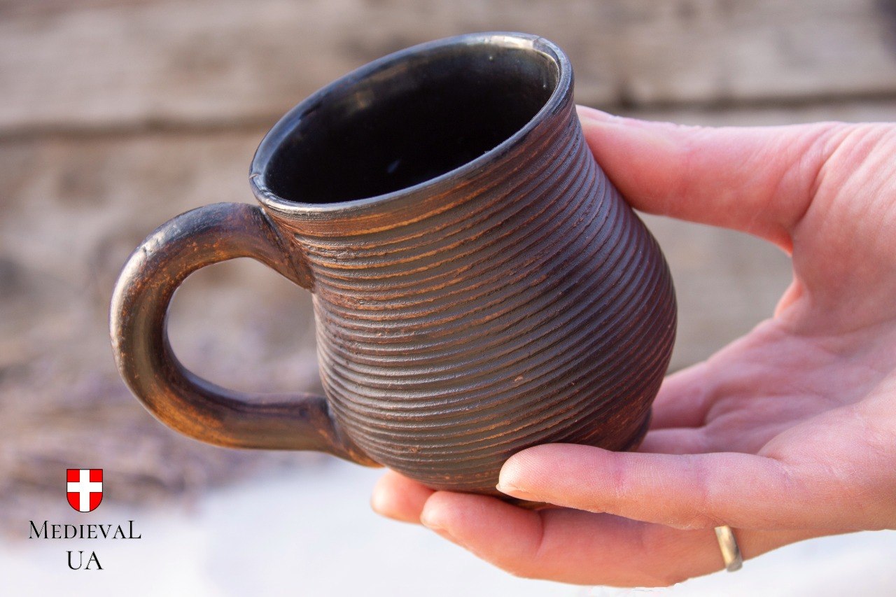 Hand cupping the small ribbed dark milk-fired medieval coffee mug, showing its rounded belly
