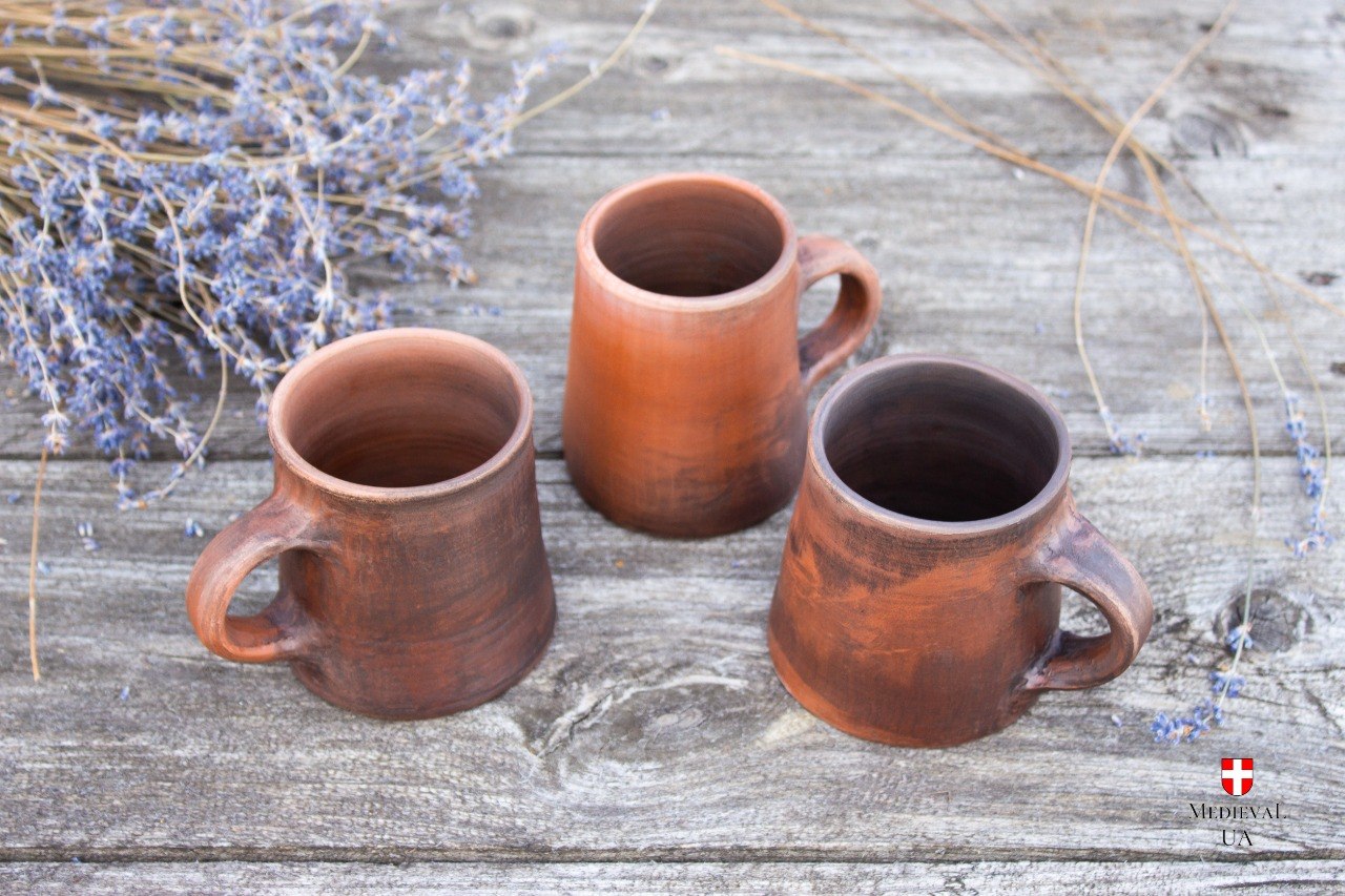 Three handmade red clay medieval cups on weathered wood showing natural color variation from light to dark brown