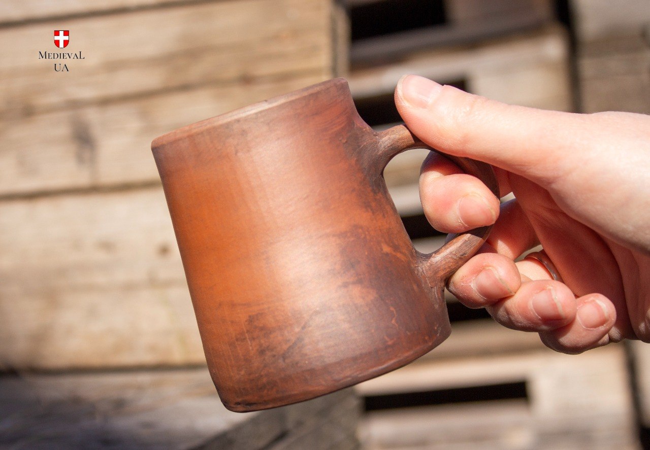 Hand gripping the red clay medieval cup by its loop handle in sunlight