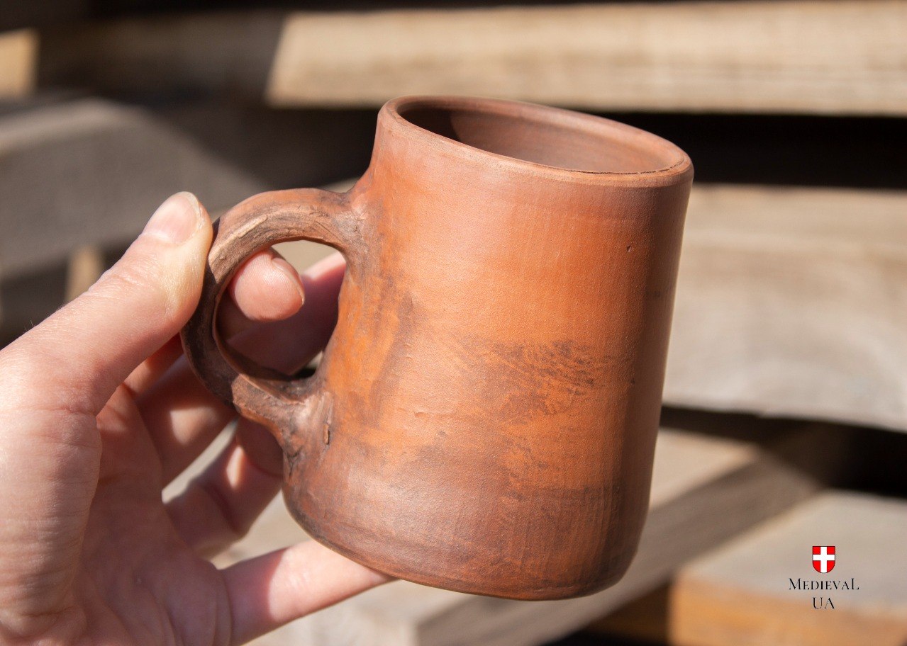 Hand holding the rustic red clay medieval mug in sunlight, showing the small handle and smooth surface