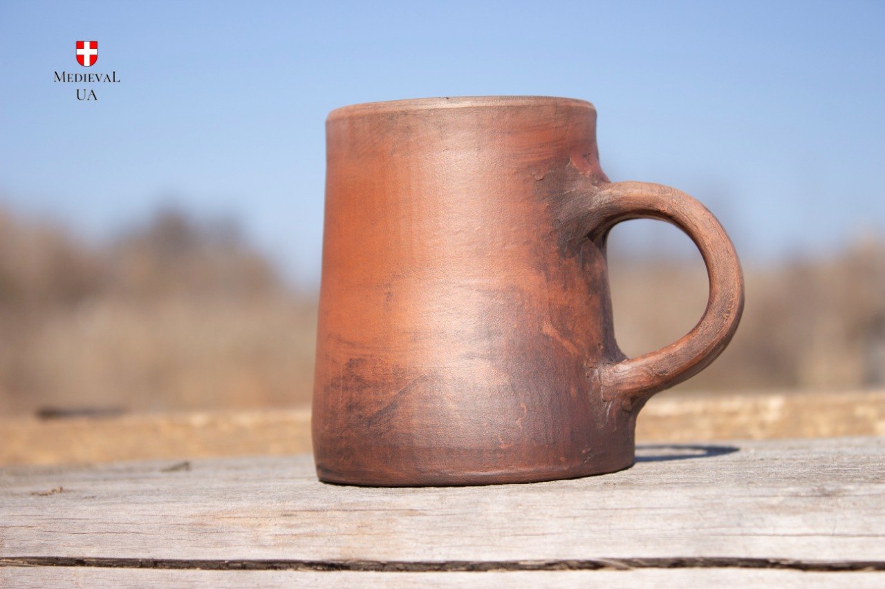 Side view of the handmade red clay medieval cup on wood against a clear sky