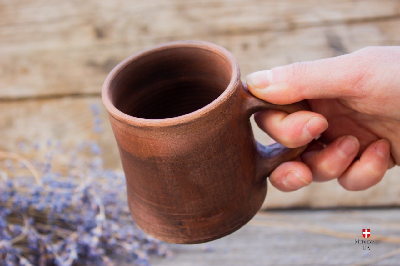 Hand holding a handmade medieval red clay cup with a small loop handle, dried lavender on weathered wood behind