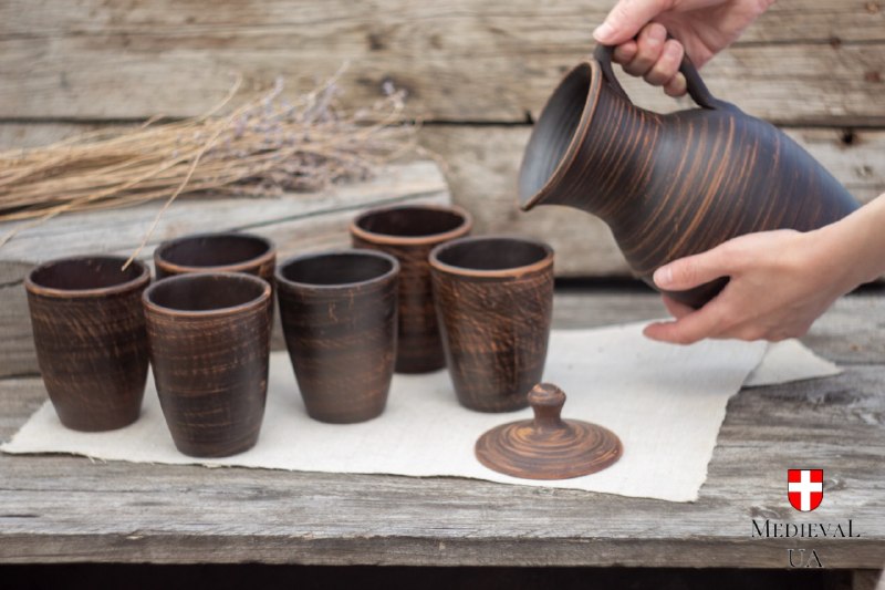 Hands pouring from a handmade clay pitcher into six matching unglazed cups on rustic wood
