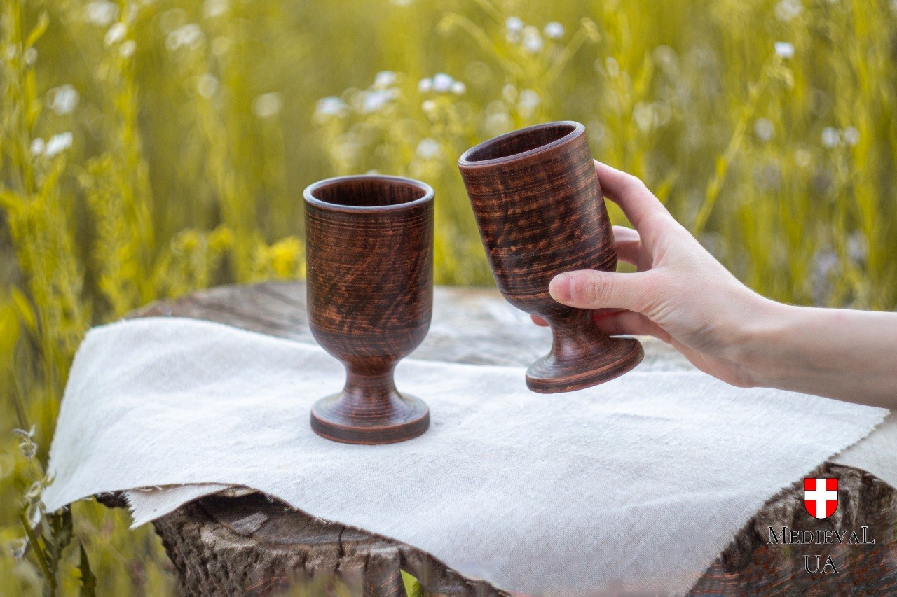 Two handmade red clay goblets on linen in a meadow, one held in hand, showing streaked milk-fired surface