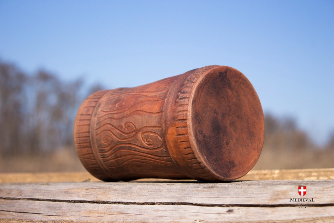 Carved red clay medieval mug lying on its side against a blue sky, showing the textured body and base