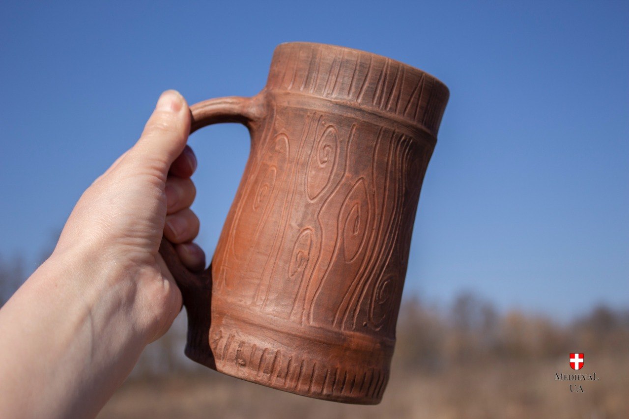 Hand raising the handmade red clay Viking tankard, highlighting the carved wood-grain surface and C-handle