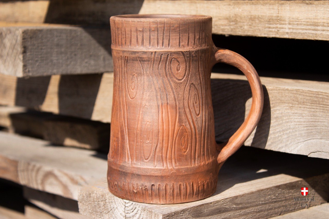 Handmade red clay medieval tavern mug with carved wood-grain texture standing in sunlight on wooden boards