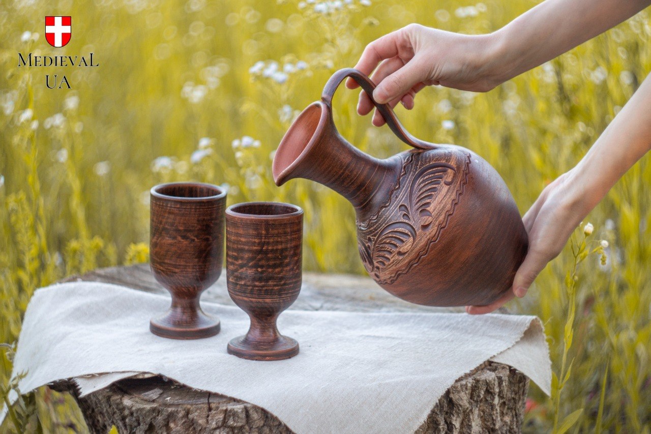 Red clay pitcher tilted to pour beside two matching goblets on a rustic stump