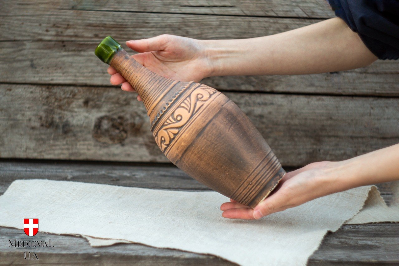 Rustic red clay mead bottle held in two hands, showing carved decorative band and green glazed neck