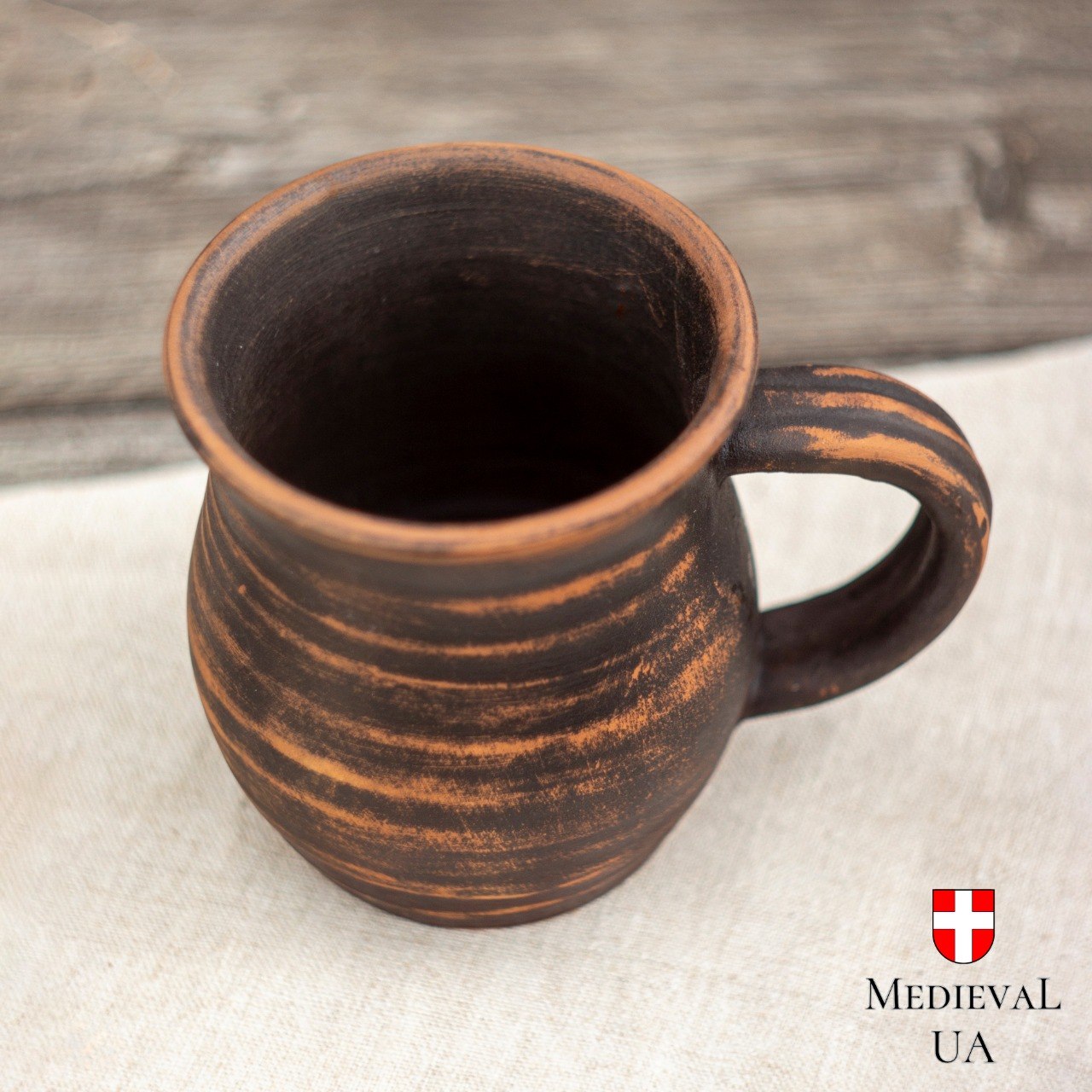 Single milk-fired red clay mug upright on linen, deep brown surface with lighter streaks and curved handle