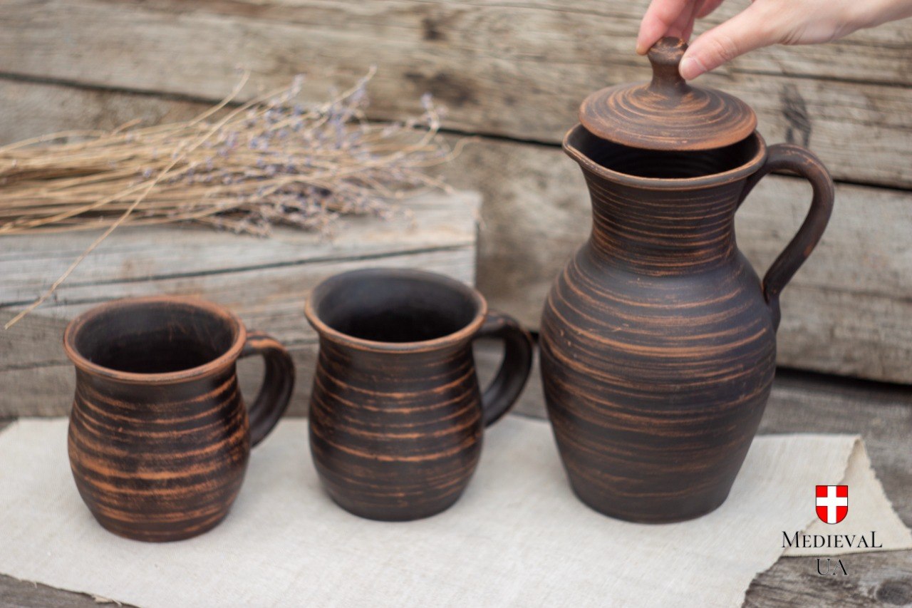 Three-piece red clay drinkware set with lidded jug and two mugs on linen and wood