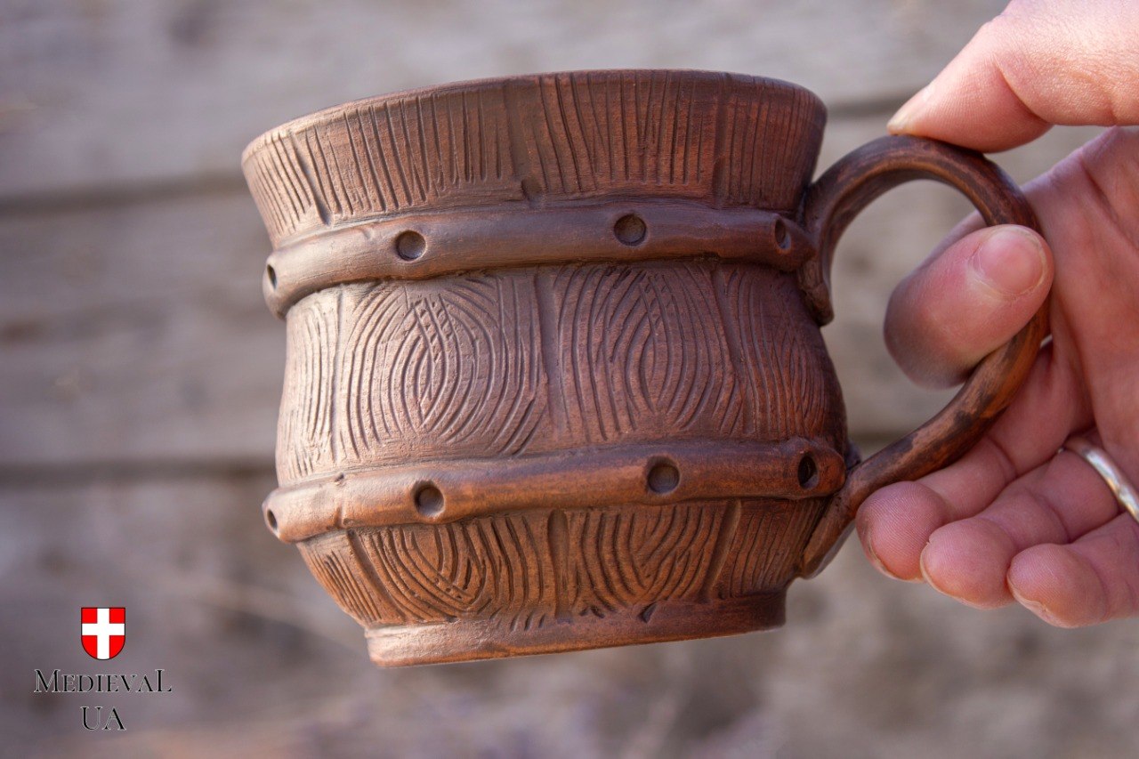 Hand holding a barrel-shaped milk-fired clay mug with carved wood-grain texture and raised hoop bands